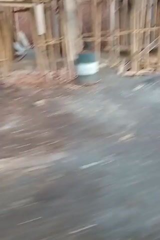 The video shows a blurry, panning view of an outdoor scene. The initial frames capture a rough, grayish surface in the foreground, with a structure resembling wooden framing or scaffolding in the background. As the camera moves, the scene transitions to reveal a circular, wooden structure supported by stilts. Further movement shows more of the surrounding environment, including some vegetation and what appears to be a rural landscape with trees and fields in the distance. The footage is shaky and out of focus throughout, suggesting it was filmed while in motion.