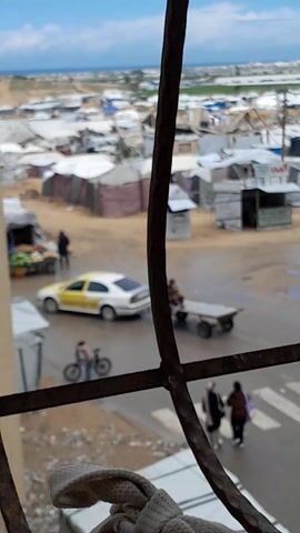 The video is taken from a window looking out onto what appears to be a refugee camp or impoverished settlement. The view is partially obstructed by a metal window frame.  Many tents or temporary shelters are visible, spread out across a sandy or dirt-covered landscape. There is a road wet from a recent rain.  A fruit stand is visible with a variety of fruits and vegetables displayed. A man is standing at the stand. A child walks with a bicycle. A moped is attached to a cart next to a yellow car. Two people are on the moped. Two other individuals walk down the road, headed away from the camera. The overall atmosphere appears to be one of poverty. The sea and horizon are visible in the background.