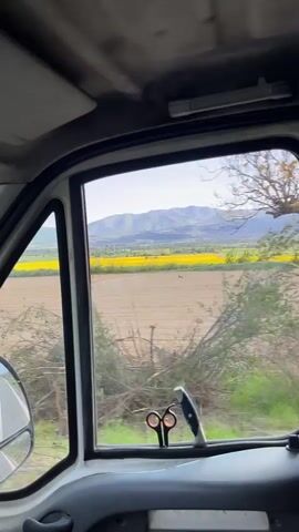 The camera is positioned inside a vehicle, likely a van, moving along a road. The view from the windows showcases a landscape of fields, with yellow patches indicating likely crops, and distant mountains. The vehicle's interior features details such as a rearview mirror with an air freshener hanging from it, an ashtray on the dashboard, and scissors and a pocket knife stuck to the window frame with tape. The road is asphalt, and the overall setting suggests a rural or countryside location.