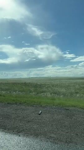 A video taken from inside a moving car shows a wide-open field of green grass under a partly cloudy sky. The car's side mirror and windshield are visible, with some raindrops on the windshield. A wiper blade moves across the windshield.