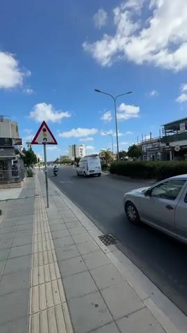 A daylight video shows a view of a street from a sidewalk. A silver sedan is driving on the street. A white van is also driving on the street. There are buildings and businesses lining the street. The sky is blue with white clouds. A traffic sign is visible on the sidewalk. The video appears to have been taken while walking or driving along the street.