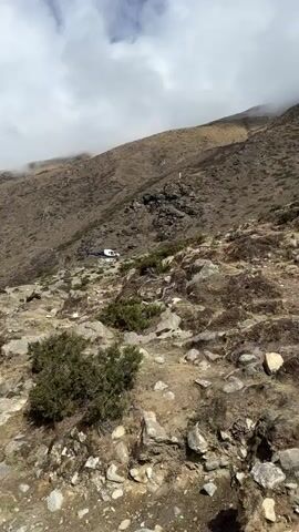 A white and blue helicopter lands on a rocky, uneven mountainside, surrounded by sparse vegetation and larger rock formations. The terrain is arid and brown, typical of a high-altitude landscape. In the background, a steep, barren mountain slope rises, partially obscured by clouds at the top. As the helicopter settles, a person, dressed in dark clothing, can be seen standing near its side. The word "MOUNTAIN" is visible in blue text on the white body of the helicopter. The sky is partly cloudy with patches of blue. The scene depicts a helicopter operation in a remote, mountainous region, likely Khumjung, Nepal, as indicated by the context.