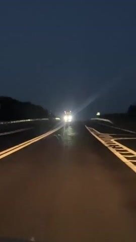 The video shows a first-person perspective of driving on a dark, wet highway at night. The headlights of an oncoming truck are visible in the distance, creating bright reflections on the wet road. The truck appears to be a large vehicle, possibly a semi-trailer truck. The highway has clear lane markings, with double yellow lines in the center and white dashed lines on the right side. The sky is dark and overcast, suggesting it may have recently rained or is about to rain. The overall atmosphere is one of travel during nighttime conditions.