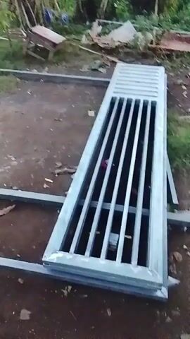 The video shows metal grates and framing on the ground. The grates appear to be for a drain or vent. The framing is made of square metal tubing. There are some plants and debris in the background. The video is shaky and the lighting is dim.