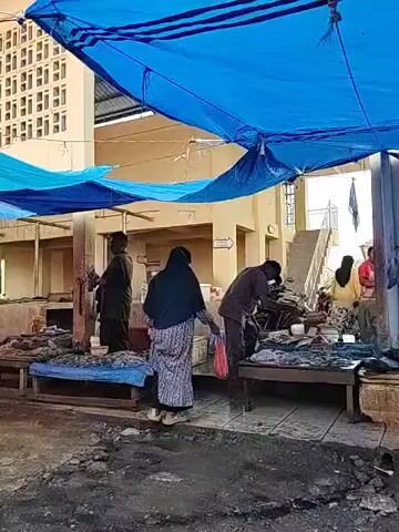 The video shows an outdoor market. People are milling about, and various vendors are selling goods. Some stalls have meat hanging. The market is covered with tarps of different colors, mainly blue and orange. The ground appears wet. The buildings surrounding the market are simple, possibly concrete structures with ventilation blocks. The video shows people interacting, examining goods, and generally going about their business in a bustling market environment.