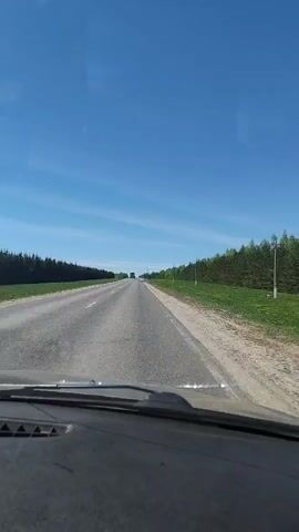 The video shows a first-person view from inside a car driving on a two-lane asphalt road. The sky is a clear blue with a few wispy clouds. The road is flanked on both sides by green fields and dense lines of trees. In the distance, several vehicles are visible on the road ahead, including a large green truck that passes by the car in the video. The dashboard of the car is visible in the foreground. The overall impression is that of a rural or countryside setting on a sunny day.