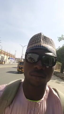 A person wearing sunglasses and a traditional hat and shirt walks down a street lined with buildings and trees. Several auto rickshaws, also known as "kekes," are seen driving on the road. The person has a backpack and a phone in their pocket. The sky is bright and sunny.