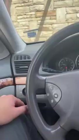 The video shows a person starting a Mercedes-Benz car. The interior of the car is visible, including the dashboard, steering wheel, and center console. The person turns the ignition, and the car starts. The background includes a stone wall and a metal structure. The camera focuses on the car's interior during the ignition process.