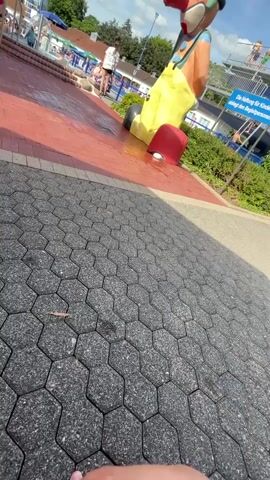 The video shows a ground-level view of a public swimming pool area on a sunny day. The foreground is paved with dark hexagonal stones. In the midground, a red tiled path leads towards a large, cartoonish statue of a character wearing yellow overalls and a red hat. The statue is positioned near a shallow pool or splash pad area where water is being sprayed. Several people are visible in the background, including children and adults, some of whom appear to be swimming or relaxing. Beyond the pool area, there are buildings and green trees. A blue sign with German text is partially visible. The camera appears to be held low, possibly by someone standing or kneeling.