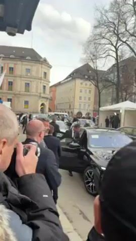 The video shows a crowded street scene in front of buildings in Munich, Germany, on an overcast day. Several black luxury sedans are parked, with people in suits exiting them. Individuals in suits and overcoats surround the vehicles, and some are seemingly providing security. A photographer is seen taking pictures. In the background, a white police van and tents are visible. Ukrainian flags are displayed on a building. The name “Lodenfrey” is displayed on a beige-colored building in the background.