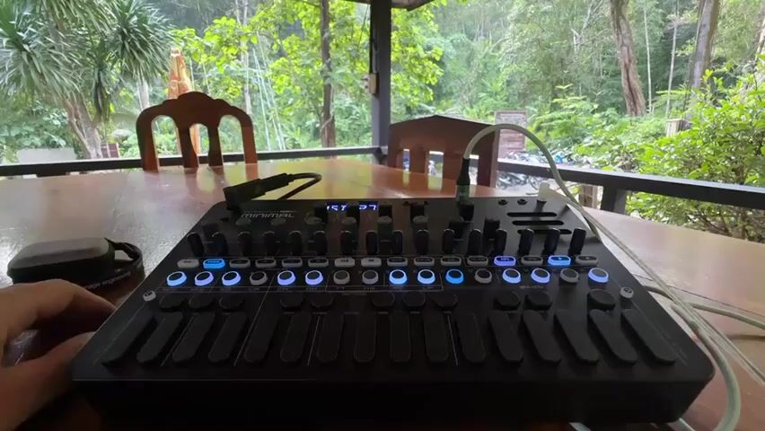 The video displays a close-up shot of a black electronic music production device, possibly a synthesizer or sampler, resting on a wooden table. The device features numerous buttons, knobs, and sliders, many of which are illuminated with blue lights. A hand is visible on the left side of the frame, near the device. Cables are connected to the device, suggesting it is powered and operational. The background reveals an outdoor setting with lush green trees and foliage, indicating the device is situated in a natural environment, possibly on a porch or patio overlooking a forest. Two wooden chairs are visible in the mid-ground, and the overall atmosphere suggests a tranquil, possibly tropical, setting. The scene is illuminated by natural light, and the camera appears to be positioned at a slightly elevated angle, looking down at the device. The video is short, consisting of a few seconds of static footage.