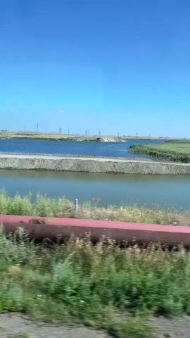 The video shows a view from a moving vehicle, likely a train, looking out over a landscape. The sky is a clear, bright blue. In the foreground, there is lush green vegetation and a large, red industrial pipe. Beyond the vegetation, there are two bodies of water, possibly ponds or shallow lakes, separated by a strip of land. The water appears calm. In the distance, there are more flat, open fields, a few scattered trees, and what appear to be power lines with tall, slender poles. The overall impression is of a rural or industrial-rural environment on a sunny day.