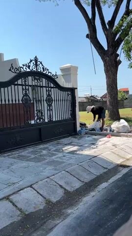 A person wearing a white hat and dark clothing is picking up trash near a large black gate and a tree. There are several trash bags on the ground. The scene appears to be outdoors in a residential area, with a house visible behind the gate and trees and buildings in the background. The ground is paved with concrete. The sky is blue and clear.