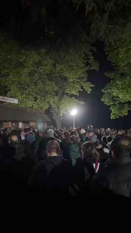The video depicts a large gathering of people at night, possibly in a park or open space. Several mature trees with green foliage are visible, and a bright light source illuminates the crowd. The people are diverse in age and attire, many wearing jackets, suggesting a cool evening. In the background, a long, low building with a tiled roof and illuminated windows can be seen. Beyond the building, a large, raised platform with bright lights suggests a stage or performance area. The camera moves slightly, panning across the crowd and showing the extent of the gathering. The overall atmosphere seems lively and potentially festive.