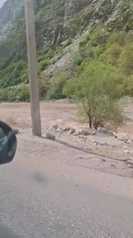 The video shows a river flowing through a mountainous region. The river is wide and the water is a muddy brown color. The banks of the river are lined with rocks and gravel. On one side of the river is a steep, rocky hillside covered in trees and shrubs. A paved road runs along the riverbank. The camera appears to be mounted in a moving vehicle, as the perspective changes slightly throughout the video. There is the sound of music in the background.