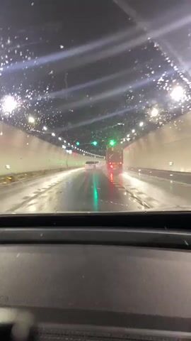 The video shows a view from inside a vehicle driving through a tunnel. The road surface is wet, and water droplets are visible on the windshield. Lights line the ceiling of the tunnel, and green arrows are displayed overhead, indicating direction. Ahead, a large truck with a container is visible, its taillights reflecting on the wet road. Another smaller vehicle is in front of the truck. The overall atmosphere is dark and wet, suggesting rainy conditions outside the tunnel.