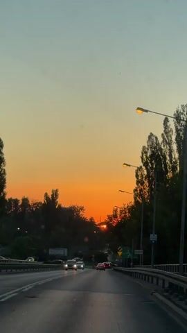 The video shows a view from a car driving on a multi-lane road, likely a highway or a major thoroughfare, as evidenced by the width of the road, the presence of multiple lanes, and the guardrails. The setting appears to be suburban or on the outskirts of a city, with trees lining the right side of the road and some buildings visible in the distance.

The most striking element of the scene is the sunset, which paints the sky in vibrant shades of orange, yellow, and a soft blue at the top. The sun itself is visible, a bright orb peeking through the trees, casting a warm glow on the landscape. The time of day is clearly sunset or dusk. The weather seems pleasant, as there are no indications of rain or harsh conditions.

Several cars are driving on the road in the same direction as the viewer. Their headlights are on, which is common during dusk or in areas with street lighting. Streetlights are also visible along the right side of the road, some of which are illuminated. The presence of traffic and streetlights suggests an urban or semi-urban environment.

On the right side of the road, there are several signs. One sign with a green background and white text is visible, which appears to be a directional road sign. Another smaller sign is also visible on a pole.

The overall mood is serene and peaceful, with the natural beauty of the sunset contrasting with the man-made elements of the road and traffic. The view evokes a sense of travel and transition, perhaps the end of a day's journey.

There is some text visible on a sign on the right side of the road. One sign has green and white lettering, typical of road signage.