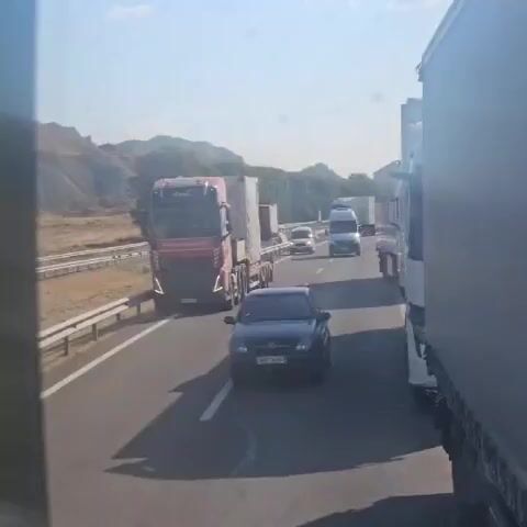The video shows a traffic jam on a highway. Several large trucks and a few cars are visible on the road. The sun is shining, and the sky is clear. The terrain on the side of the road appears to be dry and hilly. The vehicles are moving slowly or are stopped. A red truck with a white trailer is in the foreground, followed by a dark-colored car. Other trucks and vehicles are further down the road. The perspective suggests the video is being filmed from inside another vehicle, possibly a truck.