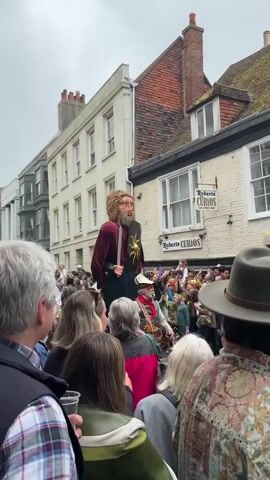 The video shows a crowd of people in a town watching a parade. A very large human-like figure is being carried through the crowd. The figure has fair hair, a long dark beard, a maroon shirt, a black skirt, and is carrying a sword in one hand and a shield with a golden sunburst design in the other. The crowd is dense and diverse, with people of all ages, including children, and wearing a variety of clothing. The parade is taking place on a street lined with buildings of different sizes and architectural styles. One of the buildings has a sign that reads "Roberts Curios." The weather appears to be overcast.