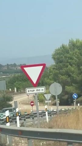 The video shows a view from a moving vehicle on a sunny day. The foreground shows a white pole and a blue and white striped traffic sign. Further in the background, there are more traffic signs and vegetation including trees and dry grass. In the far distance, there are hills and what appears to be a body of water. The sky is clear and blue. The camera appears to be shaky, suggesting the vehicle is in motion.