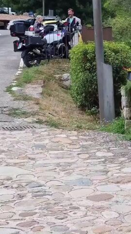 A person in a motorcycle jacket and pants stands on a cobblestone path next to a motorcycle. Another person, wearing a jacket and a backpack, is seated on the motorcycle, facing away from the camera. Behind them, a yellow car and other motorcycles are visible. The environment appears to be a rural or mountainous area with trees and greenery.