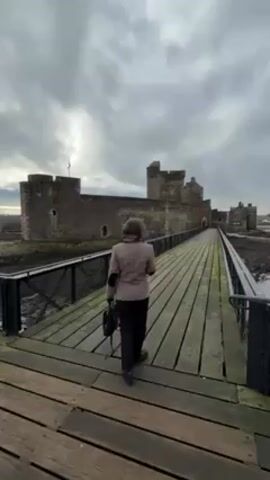 A woman walks across a wooden bridge toward a stone castle. She is wearing a tan jacket, dark pants, and a scarf around her neck, and carrying a small black backpack. She briefly turns and smiles at the camera. The bridge has black metal railings and the castle is surrounded by mud and water. The sky is overcast.