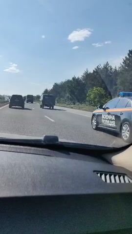 The video shows the view from inside a car, looking out onto a multi-lane highway. The day is sunny with a blue sky and scattered clouds. The road is straight and lined with trees on the right side. Several vehicles are in front of the camera car, including a dark-colored sedan, a military-style truck with "POLIS" written on the back, and a van. At one point, another light-colored van is seen passing on the right. The dashboard of the car is visible in the foreground, along with a portion of the passenger's arm and hand.