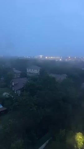 A video captures an aerial view of a neighborhood under a hazy, blue sky, likely during twilight or a storm. The scene is dominated by dense green trees interspersed with houses and a tall, multi-story apartment building. The apartment building, a prominent feature, is white with yellow accents and features numerous lit windows. The ground below the apartment building appears to be a landscaped area with pathways and lights. In the distance, a line of lights suggests a more populated area or road. The video also shows rain droplets on the camera lens, indicating current weather conditions. The overall mood is somber and somewhat obscured by the atmospheric haze and rain.