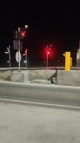 The media file captures a nighttime scene at what appears to be a toll booth or checkpoint on a road. The setting is dark, with the only significant illumination coming from artificial lights. In the foreground, a paved road is visible, marked with white lines. On the right side of the road, there are concrete structures and yellow and silver metallic barriers, typical of a toll collection point. Overhead, traffic lights are prominent, all displaying red signals, indicating a stop.

In the background, beyond the toll infrastructure, a faint horizontal line suggests a distant horizon or a structure. The sky is completely dark, confirming the nighttime setting. There is a slight blur to the image, suggesting movement, possibly from the camera or the vehicle it is mounted on.

The overall atmosphere is one of quiet waiting, possibly due to the red lights halting traffic. The presence of toll infrastructure and traffic signals firmly places the scene at a point of controlled passage on a transportation route. The clear display of red lights on the traffic signals conveys a sense of stoppage and adherence to regulations. No people are directly visible in the frames, but the infrastructure implies human activity and traffic flow. There is visible text on one of the barriers in the background, reading "METTLER TOLEDO". The location context provided, "Түндүк айланып өтүүчү жол 84 га, Kyrgyzstan," indicates this is on the Northern Bypass Road, Bishkek, Kyrgyzstan. The time of day is clearly night. No specific weather conditions are discernible from the image, though the darkness might suggest clear skies or overcast conditions without precipitation.