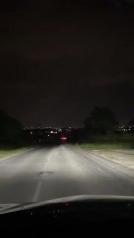 The video shows a first-person perspective of driving on a paved road at night. The headlights of the vehicle illuminate the road ahead. In the distance, there is a city or town with lights visible. The sky is dark and cloudy. There are trees and some vegetation on the sides of the road. The road appears to be a two-lane road with a white line marking the center. There are no other vehicles clearly visible on the road, though red lights are visible in the distance which could be taillights of another vehicle. The dashboard of the car is visible at the bottom of the frame. The overall scene suggests a drive through a rural or suburban area at night towards a populated area.