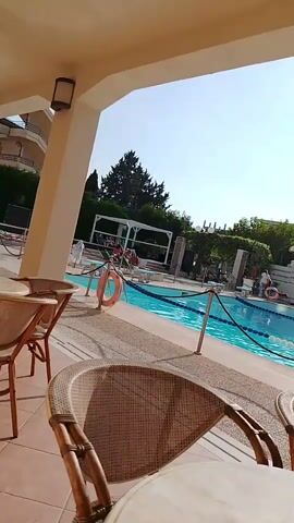 The video shows a poolside scene with tables and chairs in the foreground. A swimming pool with blue water is visible in the background, with people relaxing around it. There are lounge chairs and umbrellas around the pool. The weather appears to be sunny. On one of the tables in the foreground, there is a coffee cup and saucer, an ashtray, and some sugar packets. There are also chairs made of wicker. The scene suggests a resort or hotel with recreational facilities.