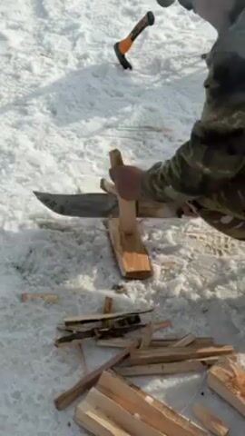 A person standing in the snow is shown cutting wood with a large knife. The person is wearing camouflage clothing and sunglasses. The snow is white and there is an axe lying nearby. The person places a small piece of wood on a larger log and cuts it with the knife. There are other pieces of cut wood scattered on the snow.