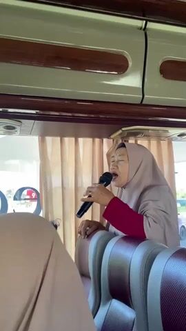 The interior of a moving bus is shown from the perspective of someone seated. Several individuals, primarily women, are visible. The first woman in the frame is wearing a beige hijab and a red top, holding a microphone and singing. The camera then pans to the left, showing other women in hijabs. The camera pans again to the right, revealing another woman in a red top holding a smartphone, seemingly taking a video. The bus's interior features light-colored walls, brown trim, and a small screen on the ceiling. Outside the window, green foliage and structures are visible, indicating the bus is in motion.