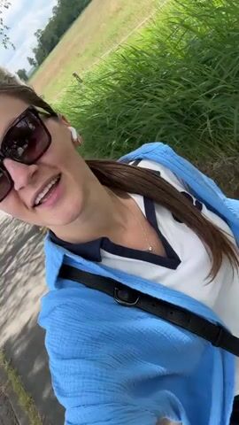 A woman in a blue shirt, white t-shirt, sunglasses, and black shorts is walking on a paved path next to a grassy field. She is smiling and gesturing with her arms as she walks.