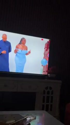 The video shows a television screen displaying a game show. Three people are on the screen: a man wearing a blue robe, a woman wearing a light blue dress, and a woman with red hair holding a light blue balloon. The woman in the dress is gesturing with her hands as if she is speaking. The television is sitting on a white cabinet with arched designs on the doors. The room is dark, and there is a table with items on it in the foreground. The wall behind the cabinet has vertical stripes.