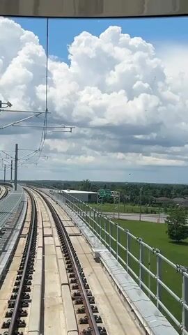 A first-person view from a train traveling along elevated train tracks. The sky is blue with large, white, puffy clouds. A silver railing runs along the side of the tracks. In the distance, there are trees, buildings, and a highway with a passing car. The train tracks curve to the left.