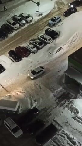 The video shows a snowy parking lot at night from an elevated perspective. Several parked cars are visible, along with a white car driving in a snowy area that could be considered a sidewalk. A pedestrian is seen walking in the upper left of the frame. The lighting is from overhead streetlights.