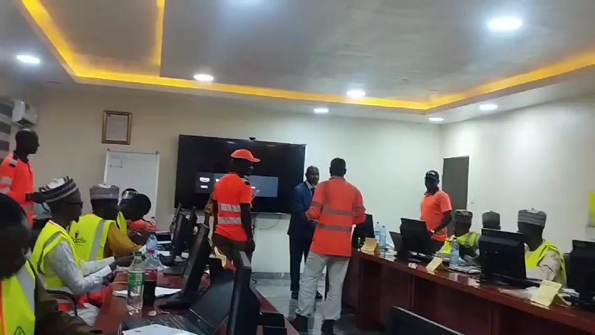 Here's a detailed description of the video:

**Scene:** The video shows a meeting room or training room.

**People:** There are approximately 10-15 individuals in the room. Most of them are seated at a long wooden table with computers. Many are wearing yellow or orange high-visibility vests over their clothing, and some are also wearing traditional hats. A few individuals, also wearing orange high-visibility vests, are standing near a flat-screen TV mounted on the wall.  One man in a suit stands with them.

**Objects:**
*   A long wooden table runs through the center of the room.
*   Several computers are set up on the table, with monitors and laptops visible.
*   Bottles of water and other drinks are on the table.
*   A flat-screen TV is mounted on the wall, displaying a screen saver.
*   A whiteboard is visible on the left side of the room.
*   Fluorescent lights are in the ceiling, providing bright illumination.

**Actions:**
*   Some people are seated at the computers, appearing to be working.
*   The men in orange vests and the man in the suit are standing and interacting, possibly in a discussion or presentation. 
*   One man calls out names and someone responds.

**Interactions:** The men in orange vests and the man in the suit appear to be addressing or instructing the people seated at the computers. The seated people appear to be engaged with the task on the computers.

**Environment:** The room appears to be well-lit, air-conditioned, and modern. The walls are neutral in color. There is a certificate or framed document hanging on the wall.