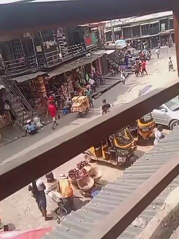 The video, shot from an elevated position, shows a busy street scene in what appears to be a market area. The view is partially obstructed by metal railings. There are people walking along the street, some carrying goods on their heads or pushing carts laden with boxes. Shops line both sides of the street. Several auto-rickshaws are parked on the side of the road. The weather appears bright, and the scene suggests a bustling commercial environment.