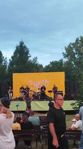 A live band performs on an outdoor stage with a yellow banner behind them that reads "С праздником славного города!" (Happy holiday, glorious city!). A group of musicians, including guitarists and a drummer, are playing on the stage. In front of the stage, an audience is seated on benches, watching the performance. The sky is overcast, and trees are visible behind the stage. The foreground shows several audience members, some of whom are looking towards the stage.