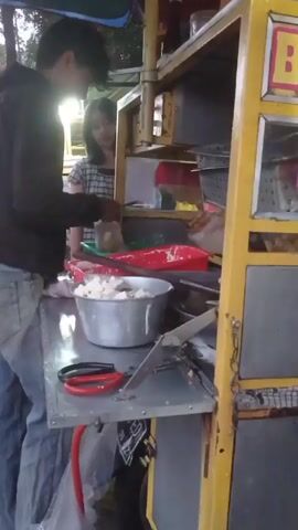 A man and a woman are standing at a food cart. The man is wearing a black hoodie and jeans. The woman is wearing a checkered shirt. The food cart is yellow and has shelves filled with various items. There is a large metal bowl on the counter of the cart, containing white food items. A pair of red-handled scissors is also on the counter. The man appears to be preparing food, and the woman is watching him. The background is out of focus but appears to be outdoors, possibly a street. The lighting suggests it is daytime.