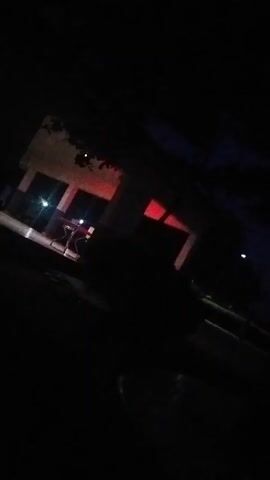 This media file depicts an outdoor scene at night in Mangu Town, Nigeria. The primary subject is a building with red light emanating from its windows, suggesting an interior light source. The building appears to be a single-story structure with visible windows and possibly some outdoor seating or furniture in front. The surrounding environment is dark, with only faint indications of trees and a pathway or road. There are also small, bright lights visible, which could be streetlights or other artificial illumination.

The overall atmosphere is somewhat mysterious due to the darkness and the red glow. The time of day is clearly night, as indicated by the deep shadows and lack of natural light. There are no explicit signs of weather conditions, but the darkness implies it is not heavily overcast or raining enough to obscure the sky. The location is identified as Mangu Town, Nigeria, but there are no specific landmarks visible to confirm this beyond the provided context.

There are no discernible people or overt activity taking place within the frame. The focus is on the building and its illuminated interior. The darkness and the selective lighting create a mood that could be interpreted as quiet, secluded, or even slightly eerie, depending on the viewer's perspective. No visible text is present in the media file.