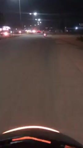The video appears to be taken from the perspective of someone riding a motorcycle or scooter at night. The road is paved and straight, illuminated by streetlights. There are other vehicles visible in the distance, including cars and another motorcycle. On the right side of the road, pedestrians are walking. The dashboard of the motorcycle or scooter is visible at the bottom of the frame. The overall environment is a suburban or urban street at night.