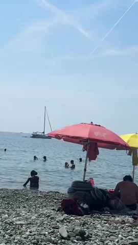 The video shows a rocky beach with people swimming in the ocean under a clear blue sky. A sailboat is docked in the distance. The camera pans to reveal a lush green mountainside behind the beach, dotted with some buildings. Umbrellas and towels are scattered on the shore, indicating a leisurely day at the beach.