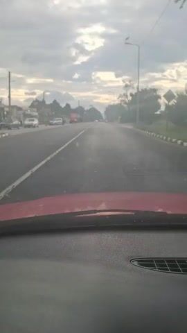 The video shows a view from the dashboard of a car driving down a paved road under a cloudy sky. The road has a double white line in the center. On the left side of the road, there are some houses and a red truck. On the right side of the road, there are trees and a streetlight with a bird on top. The car appears to be moving at a moderate speed.