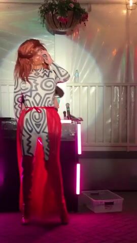 A drag performer is on a stage, wearing a white bodysuit with black tribal-like markings, a red skirt, and knee-high red boots. They have long red hair and are wearing makeup. The performer is dancing and gesturing with their hands. In the background, a person stands behind a DJ booth, and there are lights and decorations. The setting appears to be an outdoor venue, possibly a bar or club, with a tiled floor and white fencing. The lighting is dramatic, with pink and white lights illuminating the performer. The overall mood is energetic and festive.