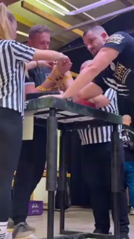 Two men are setting up to arm wrestle. The two men are standing at an arm wrestling table, with each man facing the other. Two referees, dressed in black and white striped shirts and black pants, are assisting the men with securing their grip using yellow arm wrestling straps. The room appears to be a venue, with other people milling about in the background, and artificial lighting from overhead. The men are both wearing t-shirts. The man on the left is wearing a black t-shirt with white writing. The man on the right is wearing a black t-shirt with a lion graphic. The arm wrestling table is black and gray.