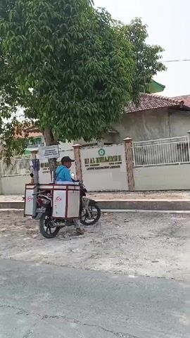 A man wearing a blue shirt and a black cap sits on a motorcycle with a sidecar. The sidecar is white with red trim and appears to be used for carrying items. The motorcycle is parked on a gravel surface next to a paved road. In the background, there is a building with a sign that reads "MI AL MUHAJIRIN II". There are trees and a fence in front of the building. Another person, a child, is visible walking near the entrance of the building. The overall scene appears to be in front of a school during the daytime.