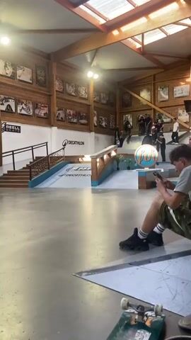 The video shows an indoor skate park. The camera focuses on a young man skateboarding. He is wearing a black tank top and black shorts. He approaches a set of stairs with a metal rail, jumps, balances on the rail, and successfully slides down it. He rolls away smoothly and stops, raising his arms in a gesture of celebration. The crowd in the background applauds. The skate park has wooden walls adorned with framed pictures. The park has a wooden beam roof and plenty of natural light. In the foreground, a person is sitting on the floor using a cell phone. A skateboard rests on the floor next to them. Logos for REELL and DECATHLON are visible on the walls.