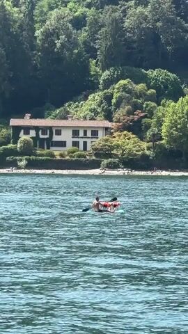 A person is paddleboarding on a body of water. The person is wearing a life vest and has a dog on the paddleboard with them. The dog is wearing a life vest as well. The paddleboarder is holding a paddle and is paddling in a direction away from the camera. In the background, there is a large house on a hill overlooking the water. The house is surrounded by trees. There are also a few other people visible on the shore in the background. The water is blue and appears to be calm. The sky is clear and the sun is shining.